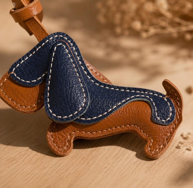 May include: A brown and navy blue leather dog-shaped keyring. The keyring has white stitching and a brown leather strap. The dog's ears and back are navy blue, while the body is brown. The keyring is on a wooden surface.
