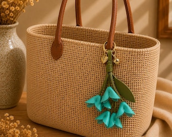Puede incluir: Bolso tote tejido beige con asas y detalles de cuero marrón. Un charm de flores turquesas cuelga del asa. El bolso está sobre una superficie de madera.