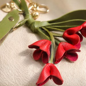 Puede incluir: Primer plano de un llavero de cuero con detalles de flores rojas. Las flores son de un rojo vibrante con un interior m&aacute;s oscuro, unidas a tallos de cuero verde y una borla de cuero verde. Los herrajes dorados completan el dise&ntilde;o.