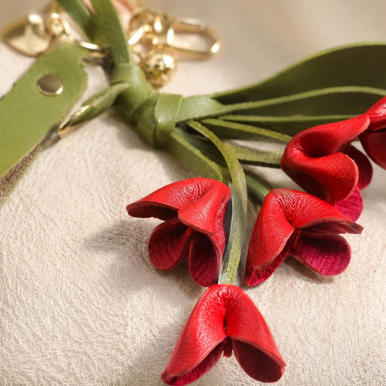 Puede incluir: Primer plano de un llavero de cuero con detalles de flores rojas. Las flores son de un rojo vibrante con un interior m&aacute;s oscuro, unidas a tallos de cuero verde y una borla de cuero verde. Los herrajes dorados completan el dise&ntilde;o.