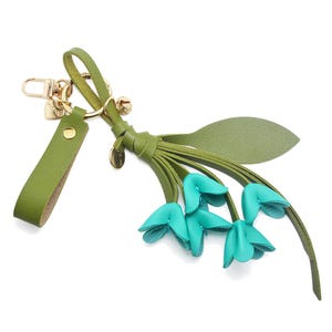 Puede incluir: Un llavero de cuero verde con cierre y herrajes dorados. El llavero presenta un grupo de flores de cuero azul turquesa y una hoja de cuero verde. Un peque&ntilde;o dije dorado cuelga del llavero.