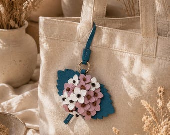 Puede incluir: Bolso tote de lona beige con un charm de flor de cuero azul. El charm presenta un racimo de flores rosas y blancas sobre una hoja azul. El bolso tiene un diseño sencillo con asas resistentes.
