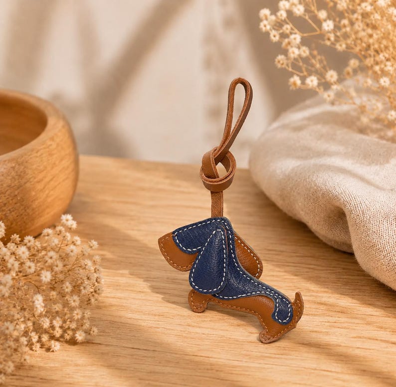 May include: A brown and navy blue leather dachshund keyring. The dog-shaped charm features white stitching and a brown leather loop for attachment. This keyring is a decorative accessory.