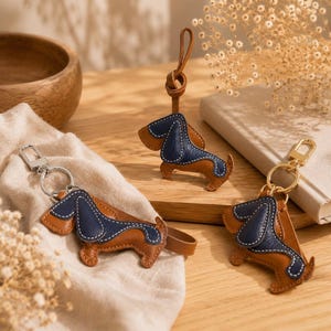 May include: Three leather dachshund keyrings. Each keyring features a brown body, a navy blue back, and white stitching. One keyring has a silver clasp, and another has a gold clasp. The third keyring has a leather loop.