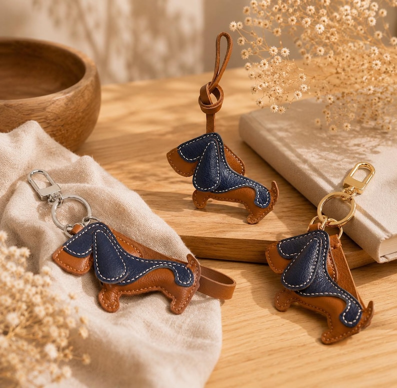 May include: Three leather dachshund keyrings. Each keyring features a brown body, a navy blue back, and white stitching. One keyring has a silver clasp, and another has a gold clasp. The third keyring has a leather loop.