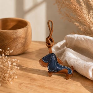 May include: A leather dachshund keyring with brown and navy blue sections, stitched with white thread. The keyring has a brown leather loop for attachment. The dog is posed on a wooden surface.