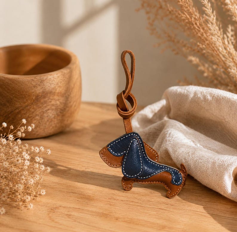 May include: A leather dachshund keyring with brown and navy blue sections, stitched with white thread. The keyring has a brown leather loop for attachment. The dog is posed on a wooden surface.