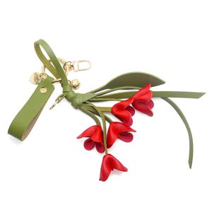 Puede incluir: Un llavero de cuero verde con un cierre dorado y un grupo de acentos de flores rojas. El llavero tiene una correa de cuero verde y un peque&ntilde;o dije dorado. Las flores son de color rojo vibrante.