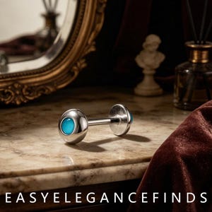 Könnte beinhalten: Ein silberfarbenes Barbell-Piercing mit einem glatten, polierten Metallschaft und zwei runden Enden, die jeweils mit einem leuchtend türkisfarbenen, blickdichten Stein besetzt sind. Der Text EASYELEGANCEFINDS ist unten aufgedruckt.