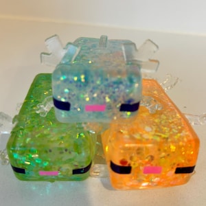 Op de afbeelding: Drie blokvormige axolotl-beeldjes gemaakt van doorschijnende hars met ingebedde glitters. De figuren zijn blauw, groen en oranje, elk met kleine doorzichtige ledematen, donkere rechthoekige ogen en een kleine roze horizontale mondlijn.