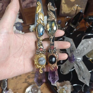 May include: Two decorative knives with black blades and ornate handles. The handles feature gold-coloured metalwork, gemstones, and amethyst crystal points. One knife has a gold leaf design, the other a swan.