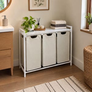 May include: A white metal laundry sorter featuring three removable beige fabric bags with oval cutouts for handles. The unit has a flat top surface and a sturdy frame, providing a structured storage solution for organizing laundry items efficiently.