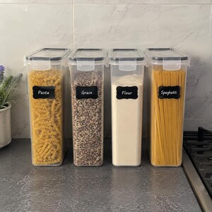 May include: Four tall, rectangular, clear plastic food storage containers with black lids and locking clips. Each container features a black decorative label with white text reading Pasta, Grain, Flour, and Spaghetti, filled with their respective contents.