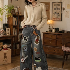 Op de afbeelding: Een vrouw draagt een beige hoodie met ritssluiting en een jeans met wijde pijpen. De jeans is versierd met verschillende geborduurde patches, waaronder teddyberen, een koffiekopje en andere grillige ontwerpen. Een bruine riem en een teddybeer sleutelhanger maken de look compleet.