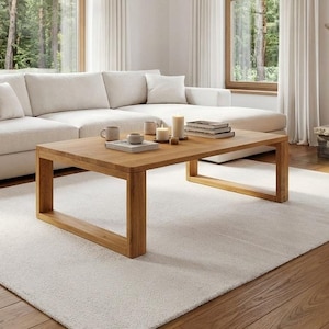 Può includere: Un moderno soggiorno con un divano angolare color crema, un tavolino in legno e un tappeto bianco. Il tavolino espone libri, candele e tazze. Sono visibili anche un camino in pietra e ampie finestre con vista sugli alberi.