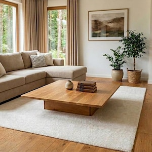Peut inclure: Salon moderne avec un canapé d'angle beige, une table basse carrée en bois et un tapis blanc. La pièce comprend de grandes fenêtres avec des rideaux beiges, une bibliothèque et des plantes en pot, créant un espace lumineux et accueillant.