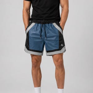 May include: Athletic shorts featuring a blue central panel, black side panels, and light gray accents. The shorts have an elastic waistband with a black drawstring and a small white emblem on the lower right side, paired with a black t-shirt.