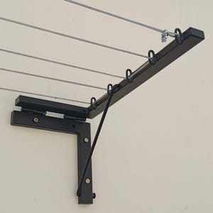 Puede incluir: Un tendedero de ropa de metal negro con múltiples líneas paralelas para colgar la ropa. El tendedero está montado en una pared y presenta un brazo plegable para soporte. El diseño incluye varios anillos de metal para colgar perchas.