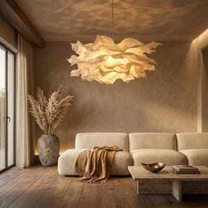 May include: A large, white, multi-layered pendant light fixture with a ruffled, cloud-like texture suspended from the ceiling. The lamp emits a soft, warm glow, casting gentle, wave-like shadows on the surrounding walls and ceiling surfaces.