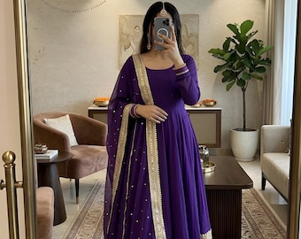 May include: A long, flowing purple dress with gold trim and a matching sheer scarf. The dress features a scoop neckline and long sleeves. The image also shows the text "VASTRA HOUSE CO."