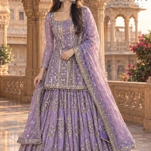 May include: A lavender lehenga choli set featuring a square-neck peplum top and a voluminous pleated skirt, both heavily embellished with gold embroidery, sequins, and mirror work. A matching sheer dupatta with a decorative border is draped over one arm.
