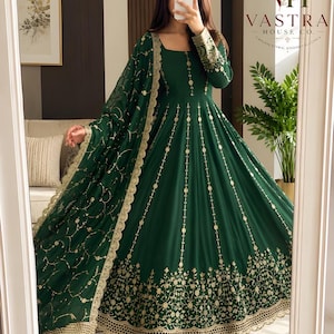 Puede incluir: Un vestido Anarkali de color verde oscuro hasta el suelo con escote cuadrado, mangas largas con bordados dorados y una falda acampanada adornada con detalles verticales de monedas doradas y un borde floral dorado en el dobladillo.