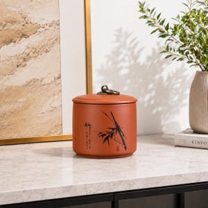 May include: A cylindrical terracotta-colored storage jar with a flat lid and a small metal ring handle on top. The exterior features a black ink-style illustration of bamboo stalks and vertical Chinese calligraphy characters on the front surface.