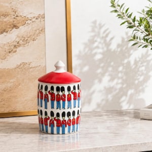 May include: A cylindrical ceramic canister with a white base and a bright red lid featuring a white finial. The exterior is decorated with a repeating pattern of British guardsmen wearing red tunics, blue trousers, and tall black bearskin hats.