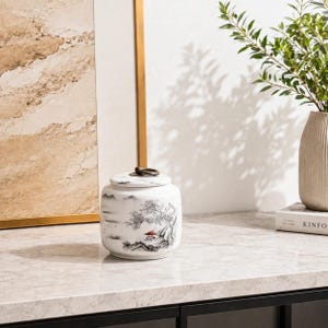 Peut inclure: Un pot en céramique blanche avec un couvercle muni d'une poignée en anneau métallique sombre. Le pot est orné d'une peinture de paysage à l'encre noire et blanche représentant des montagnes, des arbres et une petite structure rouge.
