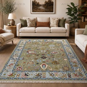May include: A large, rectangular area rug with a sage green field and a light blue border. The rug features a detailed, ornate pattern with various colors, including red, blue, and brown. The rug is placed in a living room setting with beige sofas and wooden flooring.