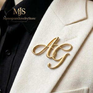 May include: A gold-toned metal brooch featuring the intertwined cursive letters AG, pinned to the lapel of a white textured blazer worn over a black collared shirt. The text MJS MonogramJewelryStore is visible in the upper left corner.