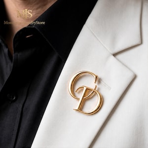 May include: A gold-colored monogram pin featuring the letters "CD" is affixed to a white blazer. The pin is a stylish accessory, adding a touch of elegance. The blazer is paired with a black shirt.