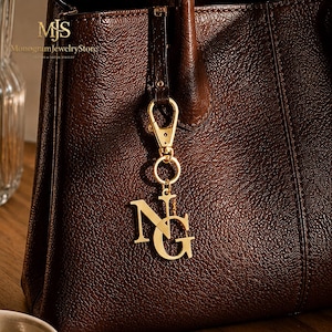 May include: A gold-toned metal charm featuring the interlocking letters N and G, attached to a gold-toned lobster clasp. The charm is suspended from the handle of a dark brown, textured leather handbag. The text MJS MonogramJewelryStore is visible.
