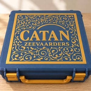 Op de afbeelding: Een blauwe, rechthoekige draagkoffer met een gouden handvat en gouden sluitingen. De bovenkant heeft een gouden rand met ingewikkelde krulmotieven en de tekst CATAN ZEEVAARDERS in grote gouden letters op het blauwe oppervlak.