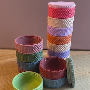 Puede incluir: Un conjunto de pastilleros circulares apilables en varios colores pastel y apagados. Cada recipiente presenta un exterior texturizado y moleteado, y está grabado con un día de la semana, incluyendo MON, TUE, WED, THU, FRI y SAT, para la organización.