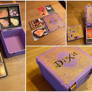 Op de afbeelding: Een paarse Dixit-spelbox met gouden accenten en de titel van het spel. De doos is open en toont spelstukken en kaarten. De kaarten liggen in een bakje en de doos staat op een houten oppervlak. Het thema van het spel is fantasie.