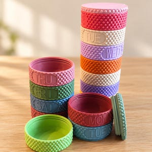 May include: A set of stackable, circular pill containers in various pastel and vibrant colors. Each container features a textured, diamond-patterned exterior and is embossed with the days of the week, including MON, TUE, WED, THU, FRI, and SAT.