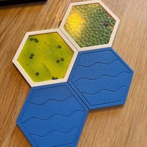 Puede incluir: Tres fichas hexagonales de juego de mesa con marcos blancos. Una ficha presenta un paisaje de bosque verde, otra muestra un campo de hierba amarilla con árboles pequeños y dos fichas son de color azul sólido con líneas onduladas.