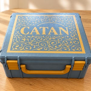 Op de afbeelding: Een blauwe rechthoekige koffer voor een bordspel met een geel handvat en twee gele sluitingen. De bovenkant heeft een goudkleurige decoratieve rand met ingewikkeld krulwerk en het woord CATAN in grote gouden hoofdletters.