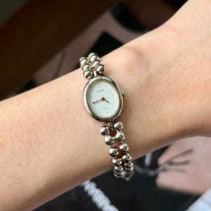 Può includere: Un orologio al quarzo ALBA color oro con quadrante ovale bianco e bracciale a perline. Il quadrante dell'orologio ha piccoli indicatori delle ore a punti. Il bracciale è composto da piccole perline rotonde. L'orologio è su un polso.