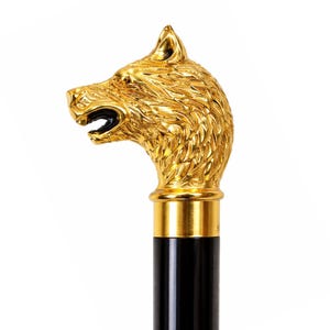 Könnte beinhalten: Ein Gehstockgriff mit einem detaillierten, goldfarbenen Wolfskopf aus Metall, der ein strukturiertes Fell und ein geöffnetes Maul aufweist. Der Wolfskopf ist auf einem glatten, zylindrischen schwarzen Schaft mit einem polierten Goldring montiert.