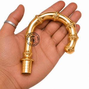 May include: A polished gold-tone metal handle shaped like a curved bamboo stalk. The handle features a textured, segmented design with a cylindrical base and a circular logo stamp that reads BRASS EMPIERS with a crown icon in the center.