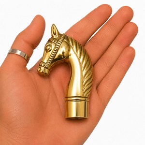 Könnte beinhalten: Ein polierter, goldfarbener Gehstockgriff aus Metall in Form eines Pferdekopfes. Das Pferd weist ein detailliertes Zaumzeug, eine glatte Mähne und einen zylindrischen Befestigungspunkt an der Basis auf. Die Oberfläche ist glänzend und metallisch.
