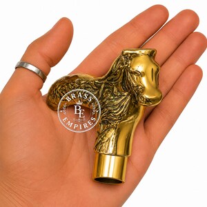 May include: A polished gold-tone metal cane handle shaped like a horse head with a textured mane. The handle features a cylindrical base for attachment and includes a circular logo with the text BRASS EMPIRES printed in white on the surface.