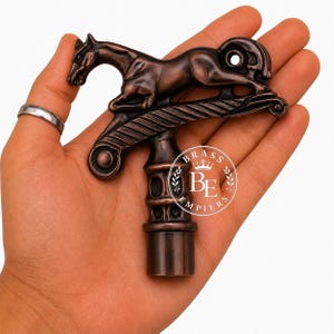 Puede incluir: Un mango de bastón de metal en tono cobre que presenta un caballo esculpido en posición de salto montado sobre una barra horizontal con una base cilíndrica. El diseño incluye un logotipo circular con el texto BRASS EMPIERS y un icono de corona.