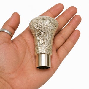 Puede incluir: Un pomo de palanca de cambios plateado con un intrincado patrón floral y de volutas en relieve en su parte superior redondeada y cuerpo cónico, acabado con una base cilíndrica de metal lisa y sencilla en la parte inferior.