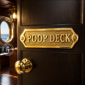 Könnte beinhalten: Ein rechteckiges Messingschild mit abgeschrägten Ecken und einem strukturierten goldenen Hintergrund, das den erhabenen Text POOP DECK in Großbuchstaben zeigt. Das Schild ist an einer dunklen Holztür mit einem passenden runden Türknauf aus Messing befestigt.