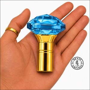 Puede incluir: Un pomo decorativo con una parte superior de cristal azul brillante tallado en forma de diamante, unido a una base cilíndrica de metal dorado pulido. El objeto se muestra sobre un fondo blanco con un logotipo circular que dice Brass Empires.