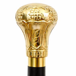 May include: A walking stick handle made of polished gold-colored metal with a rounded, bulbous top. The surface features embossed floral patterns and vertical fluting details. A smooth, gold-colored metal collar connects the handle to a black shaft.