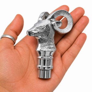 Könnte beinhalten: Ein silberfarbener Flaschenverschluss aus Metall mit einer detaillierten, dreidimensionalen Skulptur eines Widderkopfes mit markanten, geschwungenen Hörnern. Der Verschluss hat einen glatten, zylindrischen Sockel zum Einsetzen in eine Flaschenöffnung.